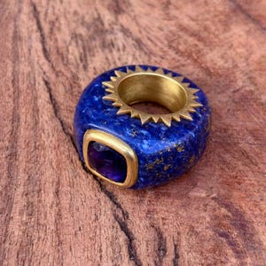 May include: A bold, blue lapis lazuli ring with a gold sunburst design and a square amethyst stone. The ring has a gold band and a polished finish, showcasing the natural beauty of the stone.