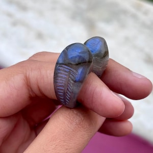 May include: Two labradorite rings with a blue and gray iridescent sheen. One ring features a carved feather design. The rings are held up by a hand. The rings are a unique piece of jewelry.