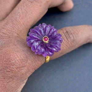 May include: A purple flower-shaped ring with a gold band. The ring features a carved amethyst flower with a small red gemstone at the center. The ring is a unique piece of jewelry.