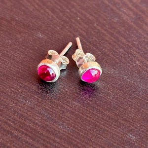 Może przedstawiać: Para srebrnych kolczyków sztyftów z okrągłymi, żywymi, magentowymi kamieniami szlachetnymi. Kolczyki mają prosty, klasyczny design, idealny do codziennego noszenia. Kamienie mają wypolerowane, błyszczące wykończenie.