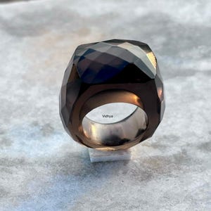 May include: A faceted, dark brown gemstone ring with a wide band. The ring has a geometric design and a polished, reflective surface. The word "Vidhya" is engraved on the inside of the ring.