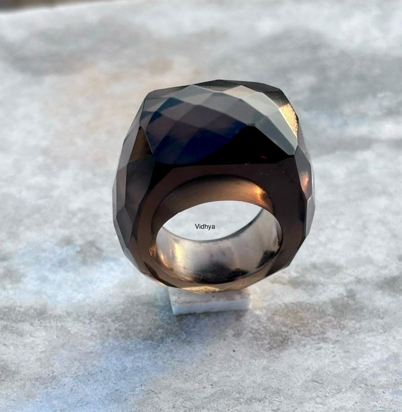May include: A faceted, dark brown gemstone ring with a wide band. The ring has a geometric design and a polished, reflective surface. The word "Vidhya" is engraved on the inside of the ring.
