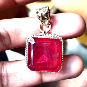 May include: A silver pendant featuring a large, square, red gemstone. The pendant has a simple, ornate design.