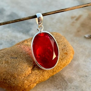 Peut inclure: Un pendentif en argent avec une grande pierre précieuse rouge de forme ovale. Le pendentif est suspendu à un cordon fin et brun.