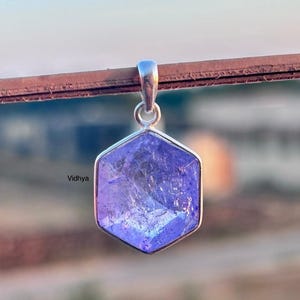 Puede incluir: Un colgante de plata con una piedra preciosa hexagonal, facetada, de color púrpura. El colgante está colgado de una cadena de plata.