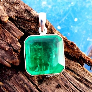 Può includere: Un ciondolo quadrato con una pietra preziosa verde smeraldo e una baionetta d'argento. Il ciondolo è appoggiato su un pezzo di legno.