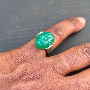 Puede incluir: Un anillo de plata con una piedra preciosa verde brillante de forma ovalada, tallada con un diseño floral. La piedra es el punto focal, engastada en una banda de plata simple y moderna. El anillo se muestra en un dedo.