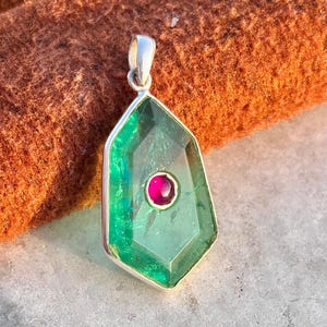 Op de afbeelding: Een edelsteen hanger met een zilveren balein. De hanger heeft een grote, facetgeslepen, smaragdgroene steen met een kleinere, ronde, robijnrode steen in het midden. De hanger heeft een zilveren rand en is geplaatst tegen een getextureerde, oranje achtergrond.