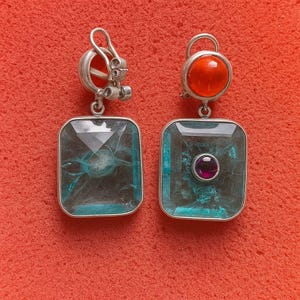 May include: Silver earrings with rectangular, teal gemstones. One has a round, orange stone at the top, the other a small, round purple stone in the centre of the teal gemstone. Set against a red background, these earrings are a striking accessory.