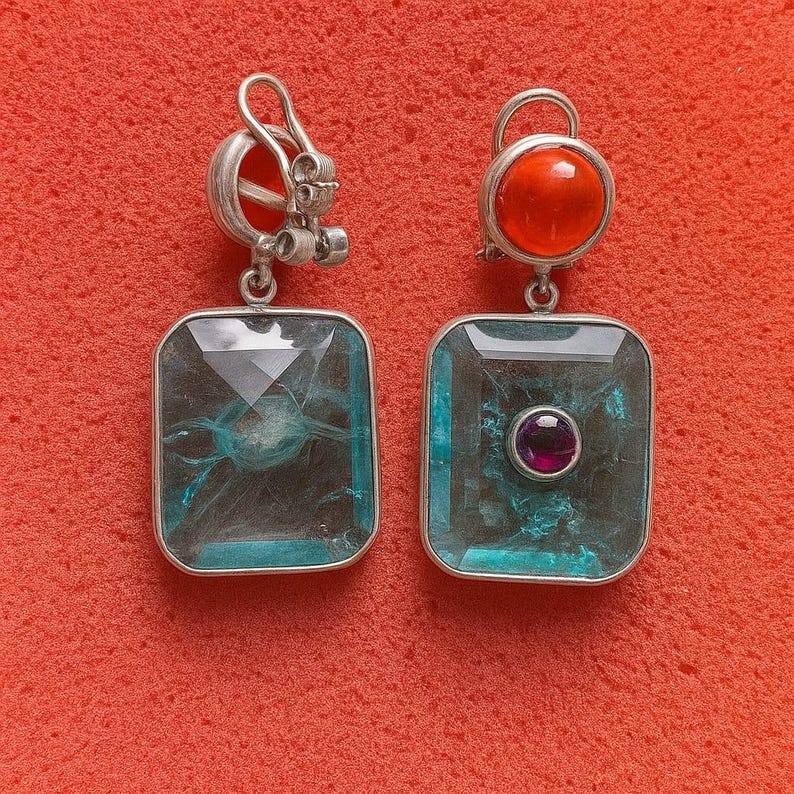 May include: Silver earrings with rectangular, teal gemstones. One has a round, orange stone at the top, the other a small, round purple stone in the centre of the teal gemstone. Set against a red background, these earrings are a striking accessory.