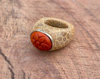 May include: A chunky ring with a beige, textured band and a vibrant orange, flower-shaped stone set in a silver bezel. The ring is displayed on a wooden surface, showcasing its unique design and color contrast.