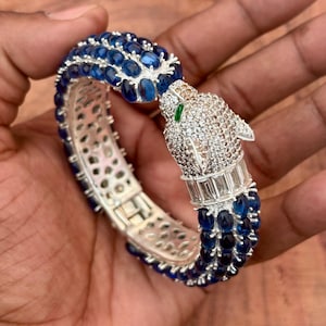 May include: A silver bracelet with a panther head design. The bracelet features blue gemstones and clear crystals. The panther's eyes are green. The bracelet is held in a hand.