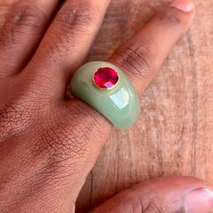 May include: A pale green ring featuring a large, round, red gemstone set in a gold-coloured bezel. The ring is on a finger, highlighting the smooth, polished surface of the stone and the ring's band.