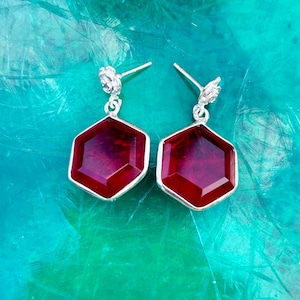 May include: A pair of silver-toned earrings with a hexagonal, deep red gemstone. The earrings have a floral design at the top and a post closure. The gemstones are set in a silver bezel.