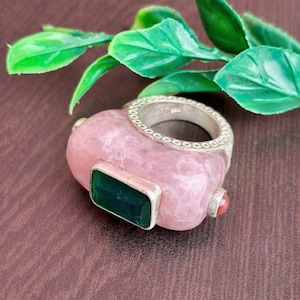 May include: A pink gemstone ring with a silver band and accents. The ring features a rectangular green stone and a small red stone. The ring is set against a brown surface with green leaves.