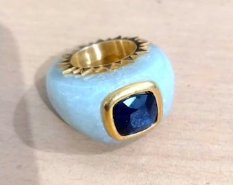Puede incluir: Un anillo con una piedra preciosa azul engastada en un engaste de oro. El anillo está hecho de cerámica blanca y tiene una banda de oro con un diseño decorativo.