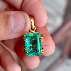 Può includere: Un pendente di smeraldo rettangolare con montatura in oro. Lo smeraldo è di un colore verde vibrante e ha un taglio sfaccettato. Il pendente ha un'ansa in oro per il fissaggio a una catena. La parola "Vidhya" è nell'angolo in basso a destra.