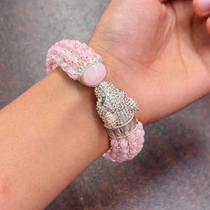 May include: A pink beaded bracelet with a silver clasp and a jeweled panther head. The bracelet features a large pink stone and small green stones for the panther's eyes. The bracelet is worn on a wrist.