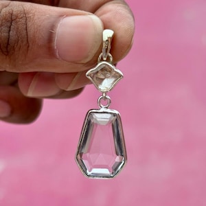 Pu&ograve; includere: Un pendente trasparente e sfaccettato con montatura color argento. Il pendente ha un design geometrico con una pietra pi&ugrave; piccola a forma di diamante sopra una pietra esagonale allungata pi&ugrave; grande. Lo sfondo &egrave; rosa pieno.