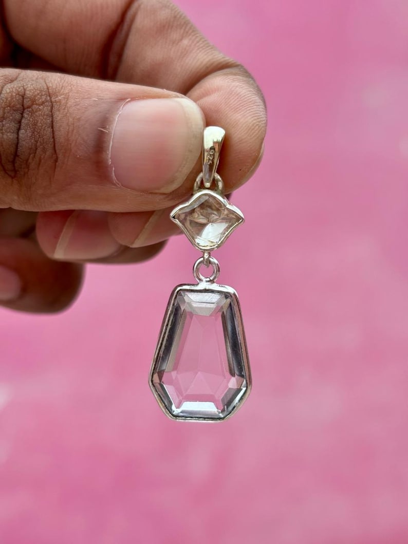 Pu&ograve; includere: Un pendente trasparente e sfaccettato con montatura color argento. Il pendente ha un design geometrico con una pietra pi&ugrave; piccola a forma di diamante sopra una pietra esagonale allungata pi&ugrave; grande. Lo sfondo &egrave; rosa pieno.