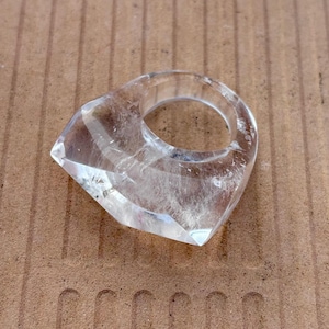 May include: A clear, faceted quartz ring. The ring has a geometric design with a large, pointed front. The transparent stone allows light to pass through, showcasing its natural clarity. This statement ring is a unique piece of jewelry.
