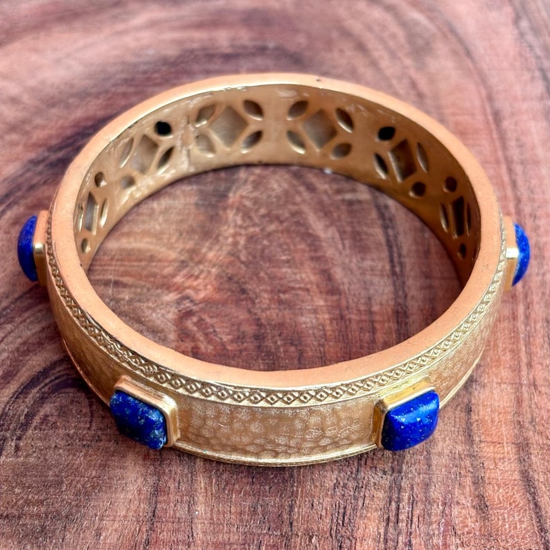 K&ouml;nnte beinhalten: Ein goldfarbenes Armreif-Armband mit drei quadratischen, tiefblauen Edelsteinen. Das Armband hat eine strukturierte Oberfl&auml;che und ein dekoratives durchbrochenes Design am oberen Rand. Das Armband wird auf einer Holzoberfl&auml;che pr&auml;sentiert.