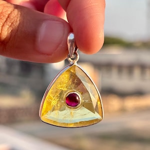 May include: A triangular pendant made of silver with a yellow gemstone and a red gemstone in the center.