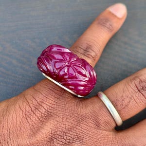 Può includere: Un grande anello di rubino scolpito con un motivo floreale. L'anello è di colore rosso intenso e poggia su una fascia d'argento. L'anello è mostrato sul dito di una persona.