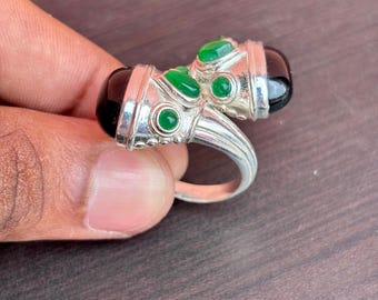 May include: A silver ring featuring two black onyx stones and several green gemstones. The ring has a unique design with a double-stone setting and decorative accents, showcasing a blend of colors and textures.