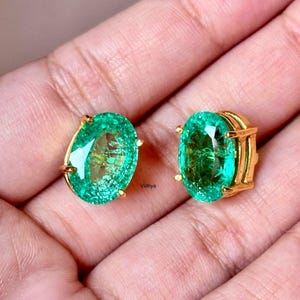 May include: A pair of oval-shaped emerald green gemstone earrings with gold settings. The earrings are set in a classic, simple style.