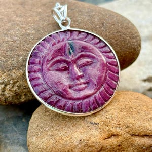 Pode incluir: Um pingente redondo esculpido com um rosto com os olhos fechados, emoldurado em uma moldura prateada. O pingente é de cor magenta escura e é suspenso por uma argola prateada. O pingente está sobre uma pedra marrom.