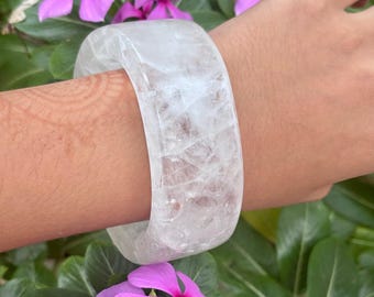 May include: A wide, translucent white quartz bangle bracelet with a smooth, polished surface and visible internal crystalline fractures, worn on a wrist against a background of vibrant pink flowers and green leaves.