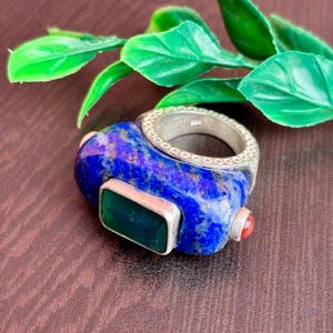 Puede incluir: Un anillo único con una gran piedra ovalada de lapislázuli y una banda de plata. El anillo tiene una piedra verde rectangular y una pequeña piedra roja. El anillo está sobre una superficie marrón con hojas verdes.
