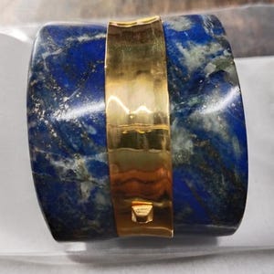 Puede incluir: Un brazalete ancho, azul y dorado hecho de lapislázuli y oro. El brazalete es una banda ancha con una banda dorada alrededor del centro.