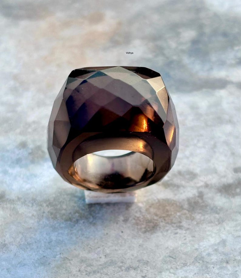 May include: A faceted, dark brown ring with a geometric design. The ring has a wide band and a large, multi-faceted stone. The ring is set on a white surface against a light gray background. The word "Vidhya" is in the top right corner.