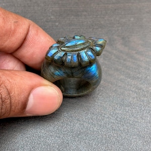 Puede incluir: Un anillo de labradorita tallada con un diseño floral. El anillo presenta una piedra central ovalada rodeada de tallas en forma de pétalos. La piedra muestra tonos iridiscentes azules y verdes, engastada en una banda gris.