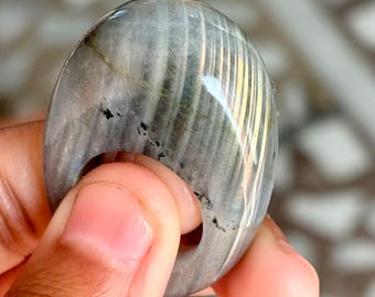 Puede incluir: Una piedra preciosa de labradorita lisa, de forma ovalada, de color gris y dorado, con un agujero perforado.