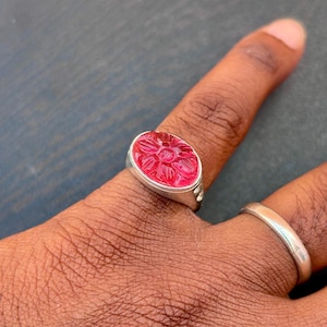 May include: A silver ring with an oval, red gemstone carved with a floral design. The ring is on a finger, and another silver band is visible on an adjacent finger. The gemstone has a detailed, three-dimensional floral pattern.