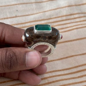 以下が含まれることがあります： 長方形のエメラルドグリーンの石をあしらった、濃い茶色の長方形のセンターピースが特徴的なユニークなリング。リングには、一連の小さな円形の切り欠きがあるシルバーバンドが付いています。小さなカラフルな石が側面にセットされています。