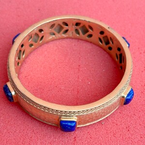 K&ouml;nnte beinhalten: Ein goldfarbenes Armreif-Armband mit quadratischen, blauen Edelsteinen. Das Armband hat ein strukturiertes Band mit einem dekorativen Muster und ausgeschnittenen geometrischen Formen. Die Edelsteine sind in goldfarbenen Fassungen gefasst.
