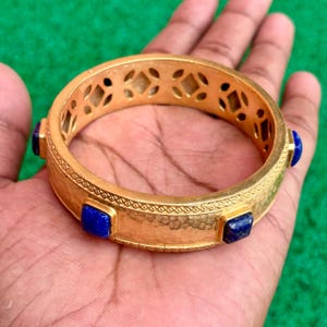 K&ouml;nnte beinhalten: Ein goldfarbenes Armreif-Armband mit strukturierter Oberfl&auml;che und geometrischen Aussparungen. Das Armband hat quadratische, tiefblaue Edelsteine, die in regelm&auml;&szlig;igen Abst&auml;nden um das Band angeordnet sind. Das Armband wird in einer Hand vor gr&uuml;nem Hintergrund gehalten.
