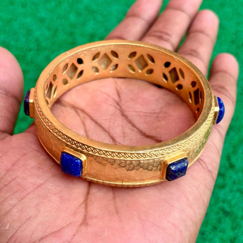 K&ouml;nnte beinhalten: Ein goldfarbenes Armreif-Armband mit strukturierter Oberfl&auml;che und geometrischen Aussparungen. Das Armband hat quadratische, tiefblaue Edelsteine, die in regelm&auml;&szlig;igen Abst&auml;nden um das Band angeordnet sind. Das Armband wird in einer Hand vor gr&uuml;nem Hintergrund gehalten.
