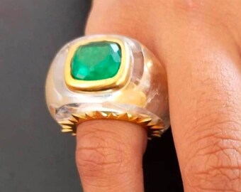 Puede incluir: Un anillo de plata y oro con una gran piedra preciosa verde esmeralda engastada en un engaste de oro. El anillo tiene una banda blanca texturizada con un borde dorado con púas.