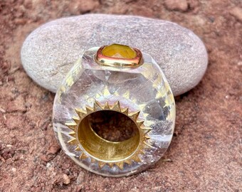 以下が含まれることがあります： ゴールドカラーのバンドと黄色の宝石が付いた、透明でファセットカットのリング。リングは、中央の開口部の周りに装飾的なゴールドカラーのサンバーストデザインが施されています。赤茶色の土の上に展示されています。