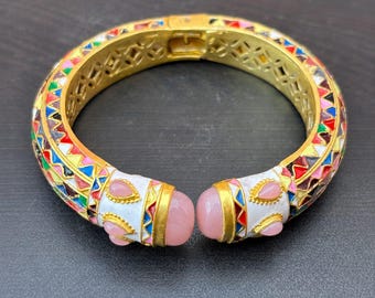 May include: A gold-tone hinged bangle bracelet featuring a repeating geometric pattern in red, blue, yellow, and white enamel. The two ends of the bracelet are finished with white enamel caps and smooth, rounded pink stones set in gold-tone metal.
