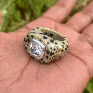 May include: A statement ring featuring a speckled, beige stone band and a square, clear gemstone set in a silver bezel. The ring's unique design is highlighted by the hand holding it.