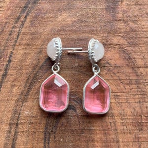 Peut inclure: Boucles d'oreilles en argent avec des pendentifs en pierres précieuses roses. Chaque boucle d'oreille comporte une petite pierre blanche ronde en haut, reliée à un pendentif géométrique rose facetté. Les boucles d'oreilles sont présentées sur une surface en bois.