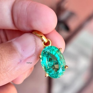 May include: A gold pendant featuring a large, oval-shaped green gemstone. The gemstone is set in a gold frame with small prongs.