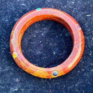 May include: A red, orange, and yellow bangle bracelet with five colorful gemstones set into the surface. The bracelet is made of a smooth, polished material.