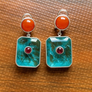 May include: Silver earrings with a striking colour scheme. Each earring has a circular, orange gemstone at the top, linked to a rectangular, teal-coloured stone with a central red gemstone. The earrings are set against a warm, brown background.